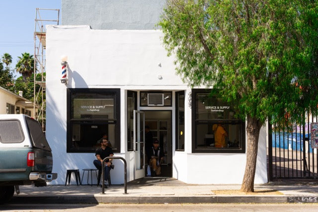White building with a person sitting outside on a chair, surrounded by trees and parked cars.